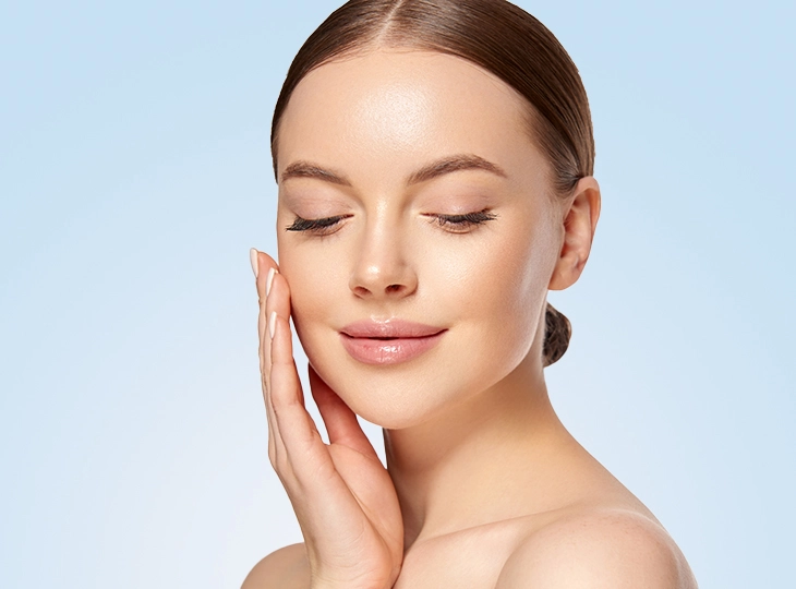 Portrait of a woman with flawless skin resting her cheek in a gentle touch, eyes closed against a light blue background.